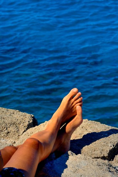 Gebräunte Unterschenkel liegen auf einer Steinmauer vor dem blauen Meer.